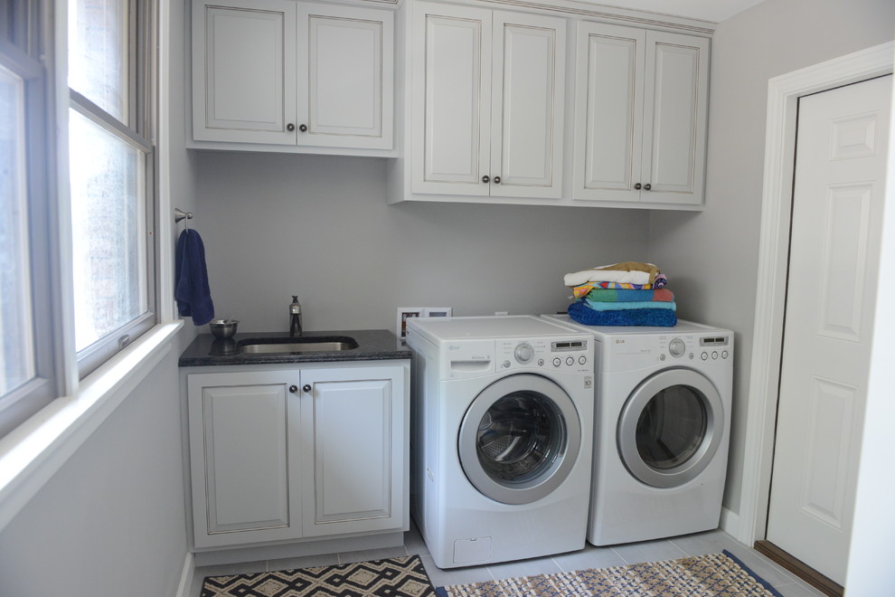 Inspiration for a mid-sized beach style single-wall utility room in Chicago with an undermount sink, raised-panel cabinets, white cabinets, granite benchtops, grey walls, ceramic floors and a side-by-side washer and dryer.