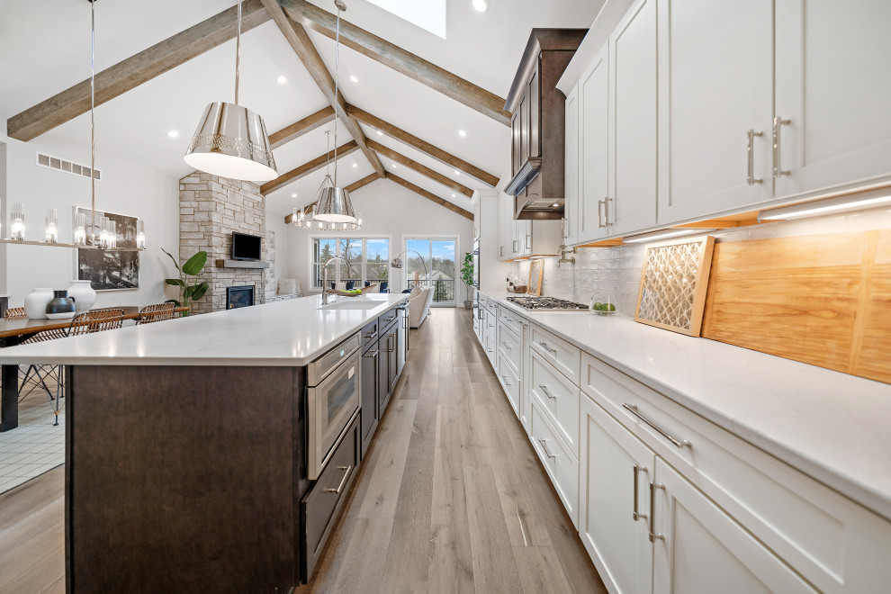 Example of a transitional galley medium tone wood floor and vaulted ceiling open concept kitchen design in Cincinnati with an undermount sink, shaker cabinets, white cabinets, quartz countertops, white backsplash, ceramic backsplash, stainless steel appliances, an island and white countertops