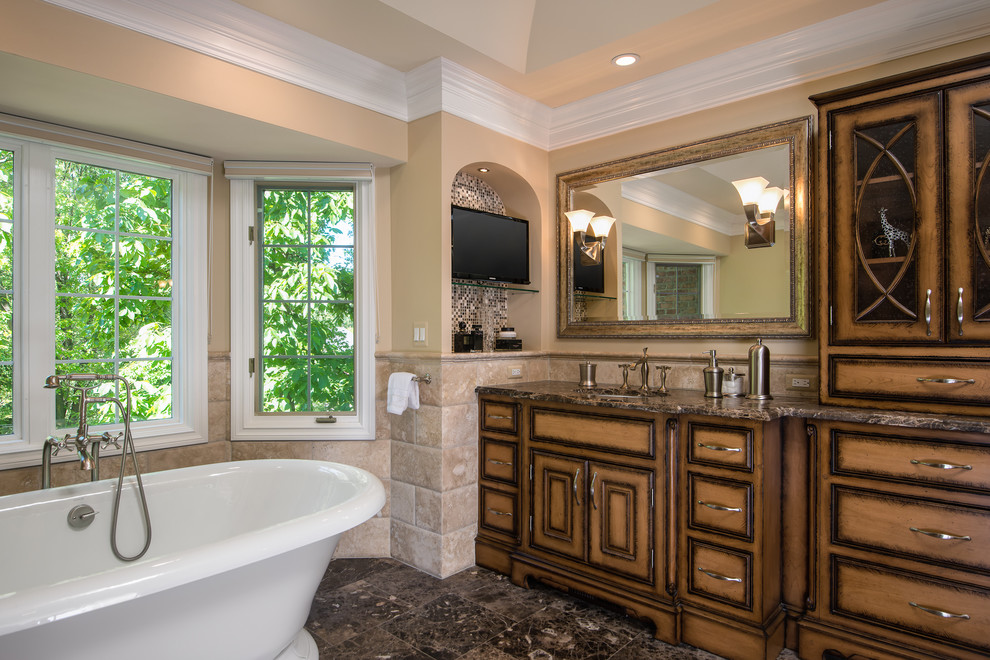 This is an example of a large traditional master bathroom in Detroit with an undermount sink, beaded inset cabinets, limestone benchtops, a freestanding tub, a corner shower, a one-piece toilet, brown tile, stone tile, beige walls, limestone floors, medium wood cabinets, brown floor and brown benchtops.