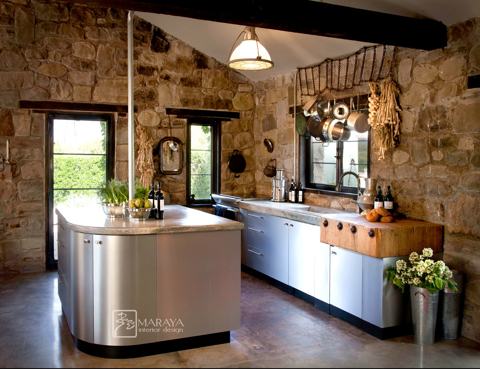 This is an example of a mid-sized country single-wall open plan kitchen in Los Angeles with a farmhouse sink, flat-panel cabinets, stainless steel benchtops, stainless steel appliances, concrete floors, with island, beige splashback, stone tile splashback and stainless steel cabinets.