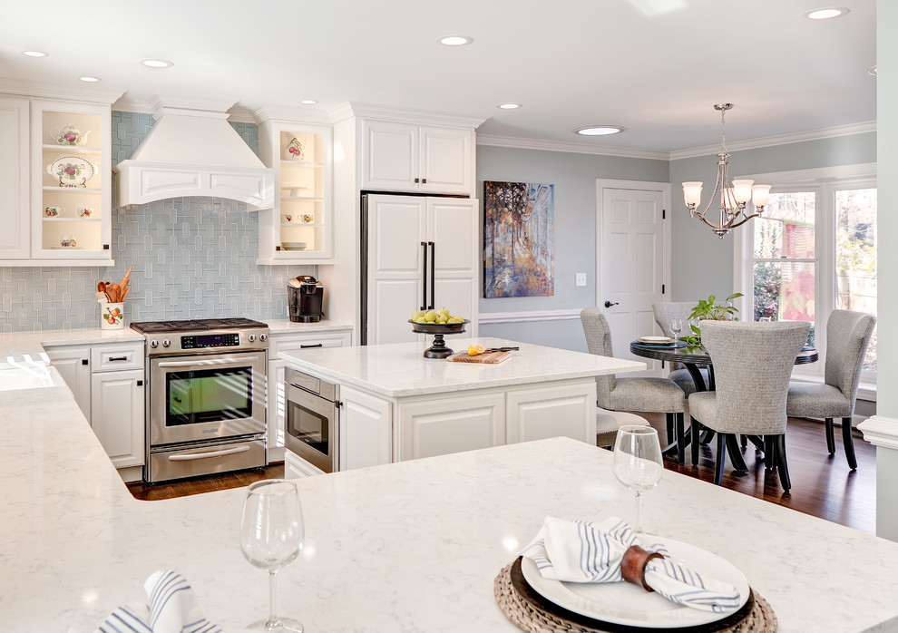 Example of a mid-sized transitional u-shaped medium tone wood floor and brown floor eat-in kitchen design with a farmhouse sink, raised-panel cabinets, white cabinets, quartz countertops, blue backsplash, porcelain backsplash, paneled appliances and an island