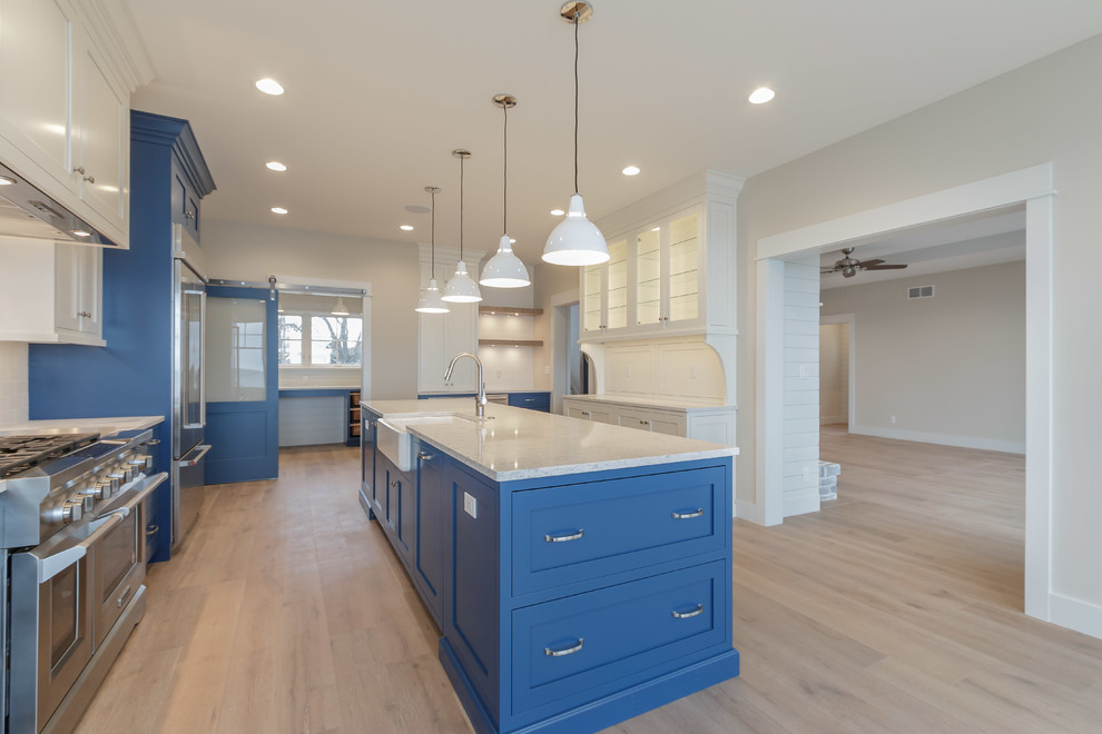 Photo of a mid-sized beach style u-shaped kitchen pantry in Other with a farmhouse sink, shaker cabinets, blue cabinets, quartzite benchtops, white splashback, ceramic splashback, stainless steel appliances, light hardwood floors and with island.