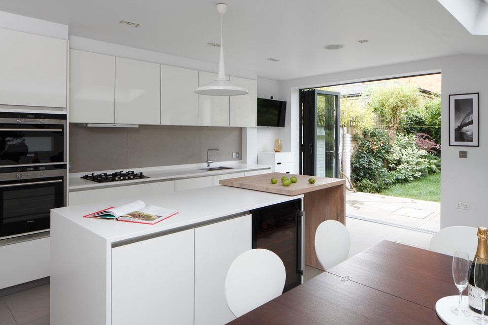 Mid-sized contemporary single-wall open plan kitchen in London with flat-panel cabinets, with island, an integrated sink, white cabinets, solid surface benchtops, grey splashback, ceramic splashback, panelled appliances, ceramic floors, grey floor, white benchtop and vaulted.
