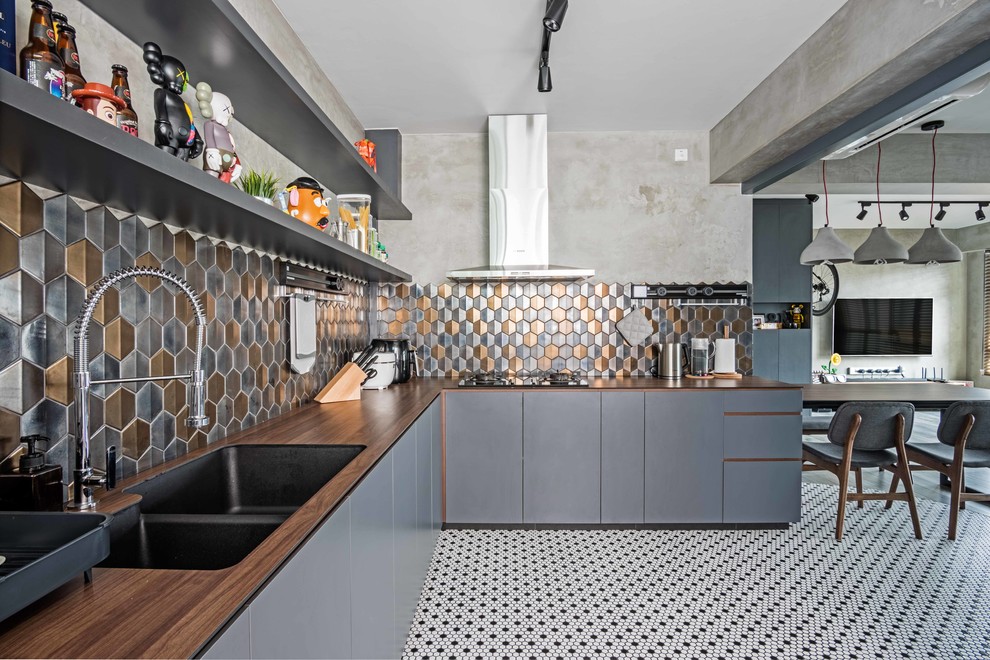 This is an example of an industrial l-shaped open plan kitchen in Singapore with a double-bowl sink, flat-panel cabinets, grey cabinets, wood benchtops, metallic splashback and multi-coloured floor.