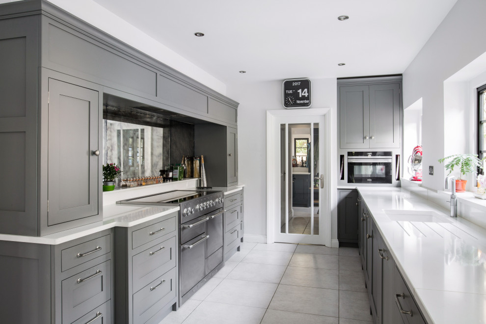 This is an example of a large transitional galley separate kitchen in Kent with an undermount sink, shaker cabinets, grey cabinets, black splashback, glass sheet splashback, panelled appliances, no island, grey floor and white benchtop.