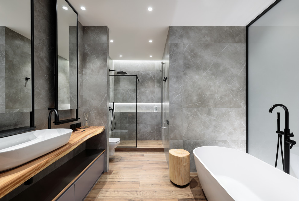 Idéer för att renovera ett mellanstort skandinaviskt brun brunt en-suite badrum, med släta luckor, grå skåp, ett fristående badkar, en dusch i en alkov, en vägghängd toalettstol, grå kakel, porslinskakel, grå väggar, ett fristående handfat, träbänkskiva, brunt golv och dusch med duschdraperi