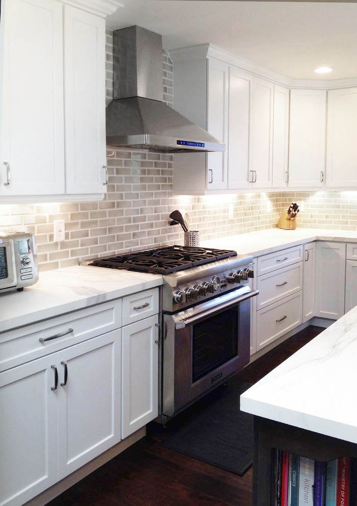 Photo of a large transitional l-shaped open plan kitchen in New York with a farmhouse sink, raised-panel cabinets, white cabinets, quartz benchtops, beige splashback, brick splashback, stainless steel appliances, dark hardwood floors, with island, brown floor, white benchtop and recessed.