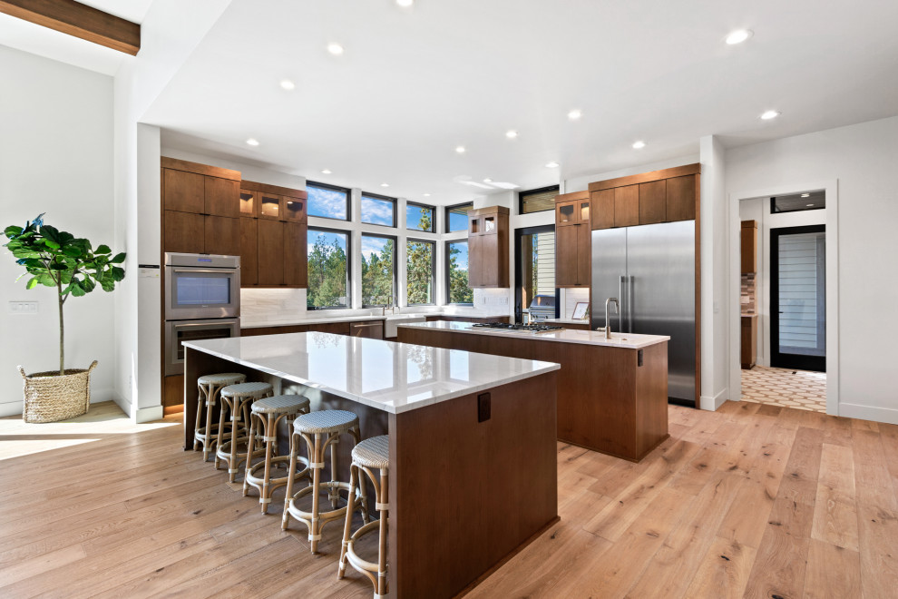 Expansive contemporary l-shaped open plan kitchen in Portland with a farmhouse sink, flat-panel cabinets, medium wood cabinets, quartzite benchtops, white splashback, ceramic splashback, stainless steel appliances, multiple islands, white benchtop, medium hardwood floors and brown floor.