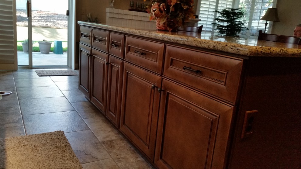 This is an example of a mid-sized traditional l-shaped eat-in kitchen in Los Angeles with an undermount sink, raised-panel cabinets, medium wood cabinets, granite benchtops, beige splashback, stone tile splashback, black appliances, porcelain floors and with island.