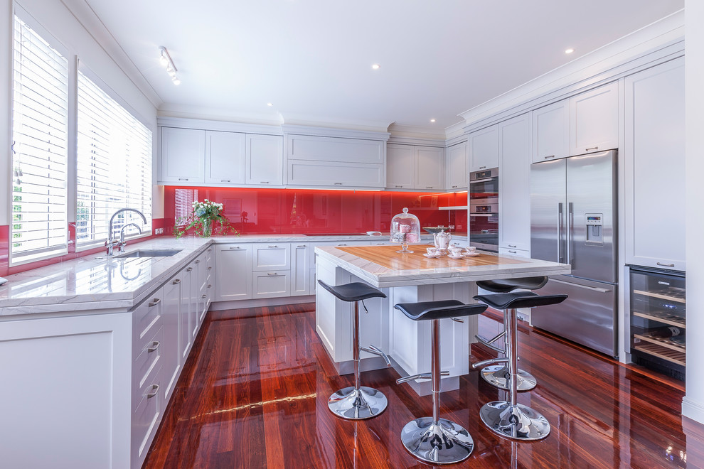 Large traditional u-shaped eat-in kitchen in Auckland with an undermount sink, recessed-panel cabinets, granite benchtops, red splashback, glass sheet splashback, stainless steel appliances, dark hardwood floors, with island, brown floor and white cabinets.