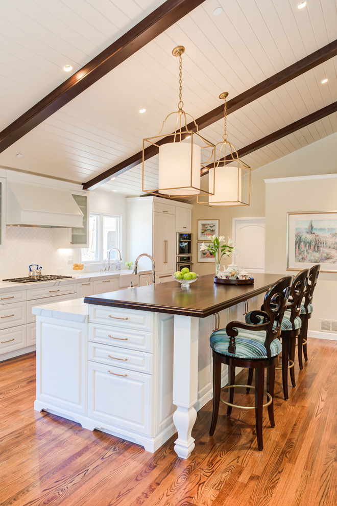 Design ideas for a large traditional l-shaped open plan kitchen in San Francisco with a farmhouse sink, raised-panel cabinets, white cabinets, wood benchtops, white splashback, ceramic splashback, panelled appliances, light hardwood floors, with island and beige floor.
