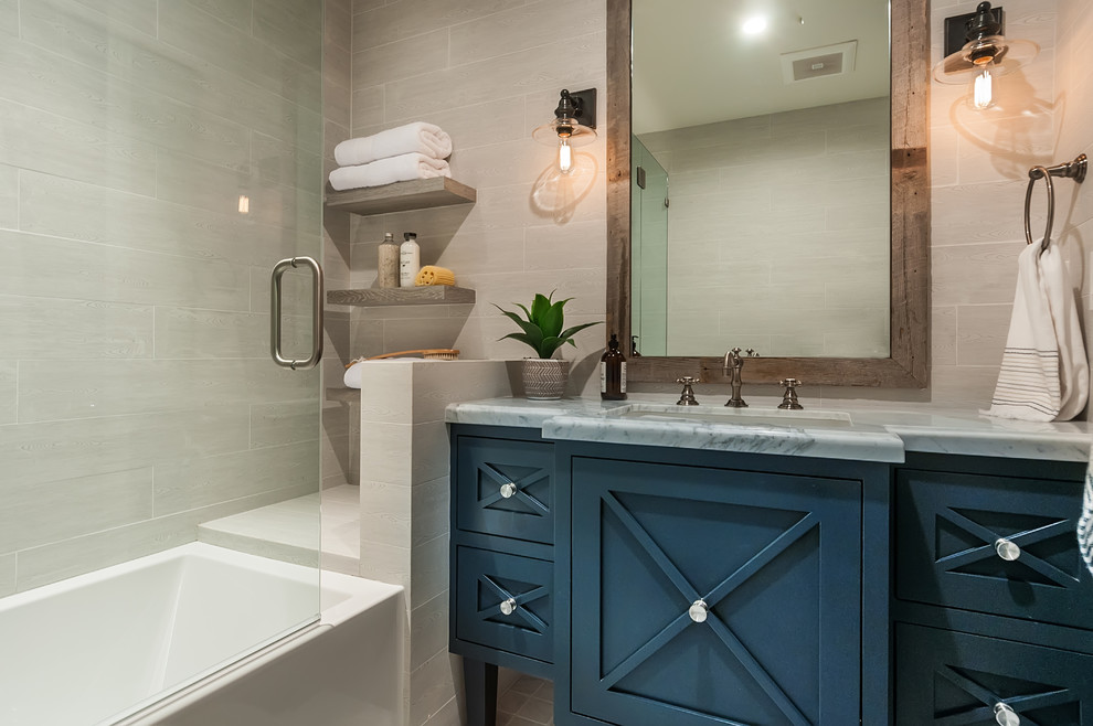 Photo of a country bathroom in San Diego with recessed-panel cabinets, blue cabinets, an alcove tub, gray tile, ceramic tile, grey walls, an undermount sink, marble benchtops, a hinged shower door and grey benchtops.