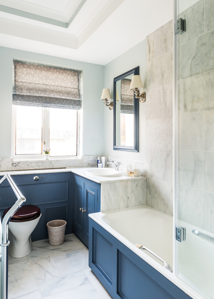 Small transitional kids bathroom in London with recessed-panel cabinets, blue cabinets, a drop-in tub, a shower/bathtub combo, a two-piece toilet, white tile, stone tile, blue walls, marble floors, a drop-in sink and marble benchtops.