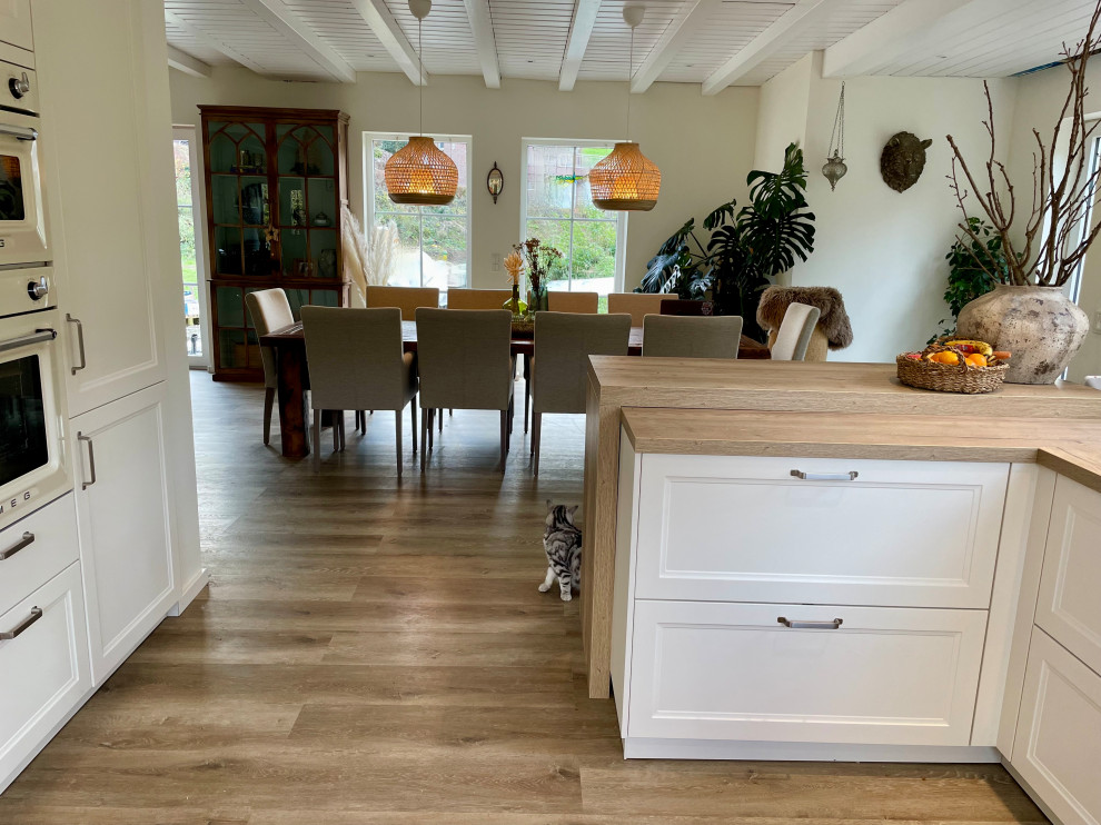 Large country u-shaped open plan kitchen in Cologne with a farmhouse sink, shaker cabinets, white cabinets, wood benchtops, beige splashback, subway tile splashback, white appliances, vinyl floors, no island, brown floor, beige benchtop and exposed beam.
