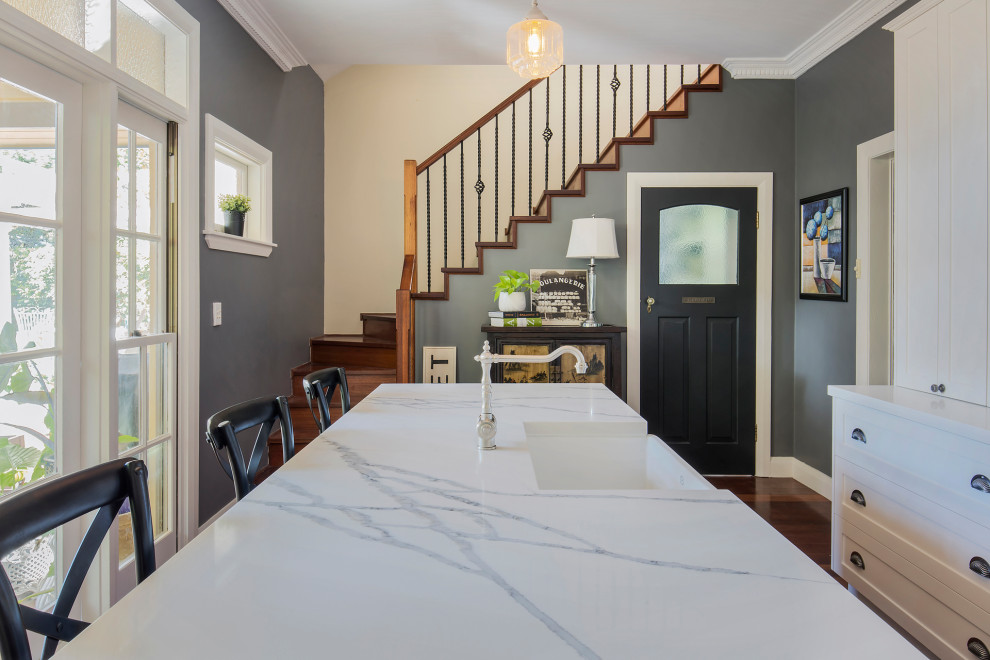 This is an example of a mid-sized traditional l-shaped eat-in kitchen in Sydney with a farmhouse sink, shaker cabinets, white cabinets, quartz benchtops, grey splashback, metal splashback, black appliances, medium hardwood floors, with island, brown floor and multi-coloured benchtop.