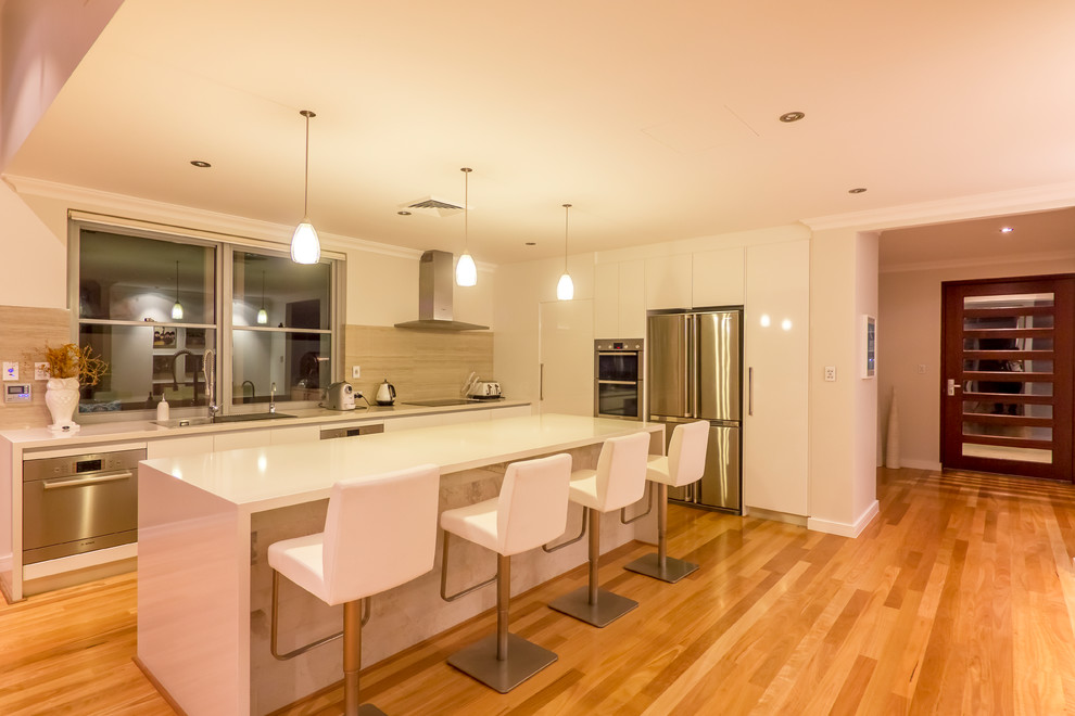 This is an example of a mid-sized beach style l-shaped open plan kitchen in Perth with a double-bowl sink, flat-panel cabinets, white cabinets, quartz benchtops, beige splashback, travertine splashback, stainless steel appliances, light hardwood floors and with island.