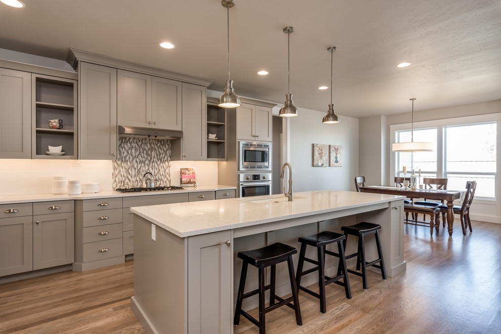 This is an example of a transitional l-shaped eat-in kitchen in Boise with an undermount sink, shaker cabinets, grey cabinets, white splashback, stainless steel appliances, medium hardwood floors, with island, brown floor and white benchtop.