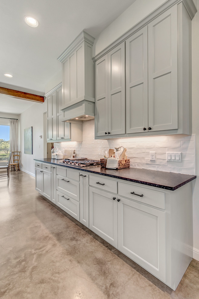 Mid-sized country galley eat-in kitchen in Dallas with a farmhouse sink, shaker cabinets, grey cabinets, white splashback, concrete floors, with island and black benchtop.