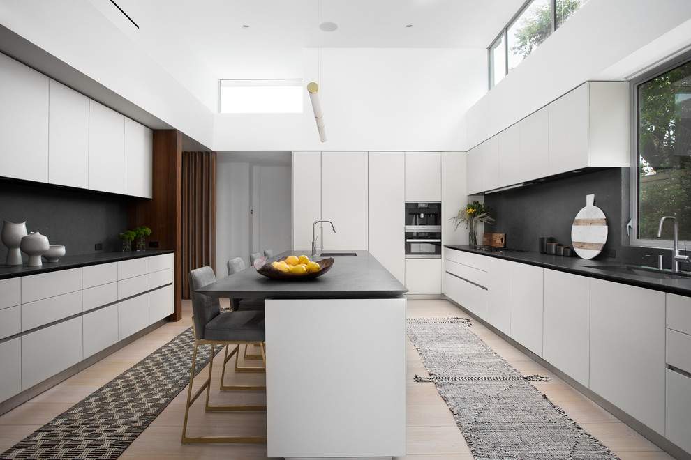 This is an example of a contemporary galley kitchen in Los Angeles with an undermount sink, flat-panel cabinets, white cabinets, black splashback, light hardwood floors, with island, beige floor and black benchtop.
