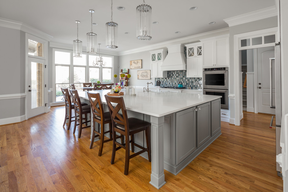 Photo of a large transitional l-shaped open plan kitchen in Raleigh with a farmhouse sink, flat-panel cabinets, white cabinets, quartz benchtops, blue splashback, glass tile splashback, stainless steel appliances, medium hardwood floors and with island.