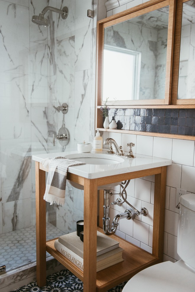 This is an example of a transitional bathroom in Minneapolis with open cabinets, medium wood cabinets, a corner shower, white tile, white walls, an undermount sink, multi-coloured floor, a hinged shower door and white benchtops.