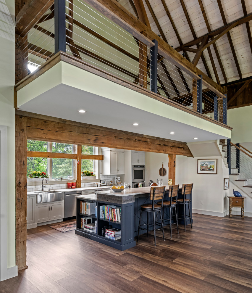 Mid-sized farmhouse galley medium tone wood floor, brown floor and exposed beam open concept kitchen photo in Philadelphia with a farmhouse sink, blue cabinets, quartz countertops, white backsplash, porcelain backsplash, stainless steel appliances, an island, white countertops and beaded inset cabinets