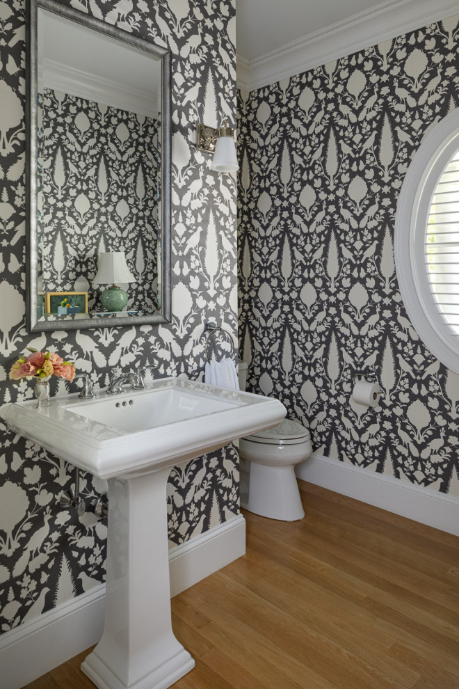 Example of a mid-sized transitional medium tone wood floor and wallpaper powder room design in Boston with white cabinets, a two-piece toilet, a pedestal sink and a freestanding vanity