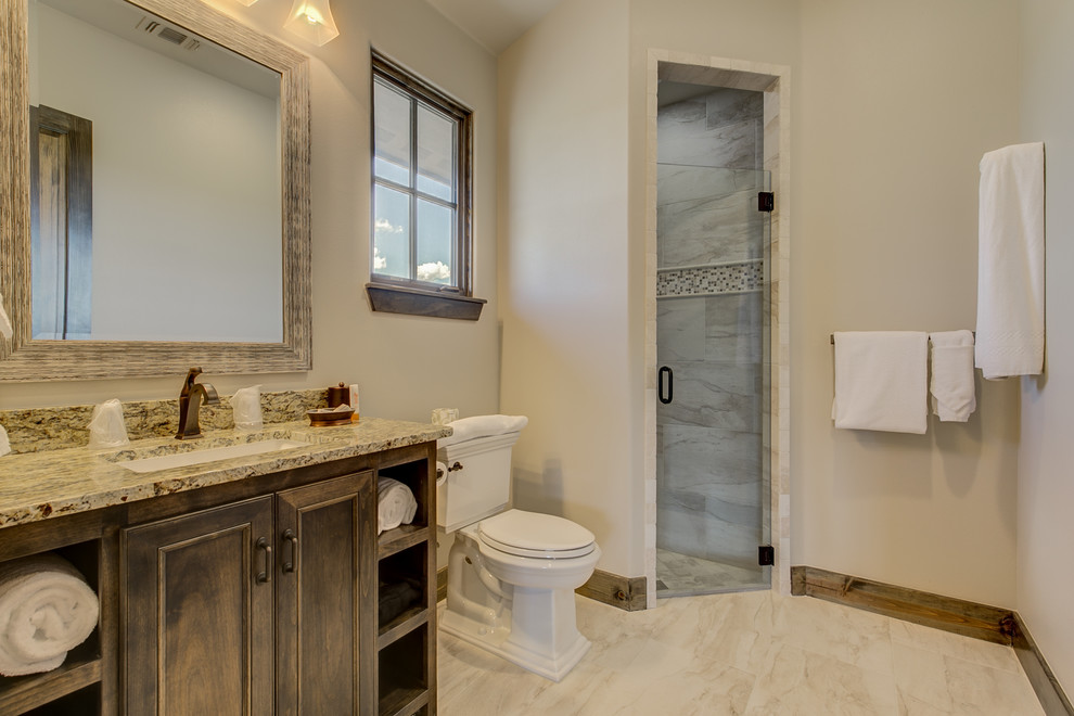 Mid-sized country 3/4 bathroom in Dallas with beaded inset cabinets, dark wood cabinets, a corner shower, a two-piece toilet, gray tile, beige walls, an undermount sink, granite benchtops, beige floor, a hinged shower door and brown benchtops.