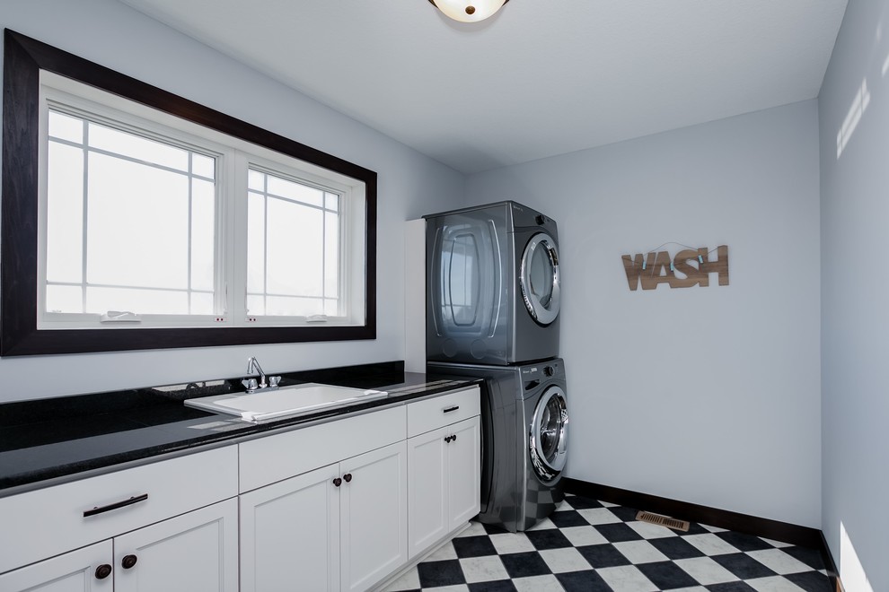 This is an example of a large transitional galley dedicated laundry room in Minneapolis with a single-bowl sink, shaker cabinets, white cabinets, solid surface benchtops, linoleum floors, a stacked washer and dryer, white walls, brown floor and black benchtop.