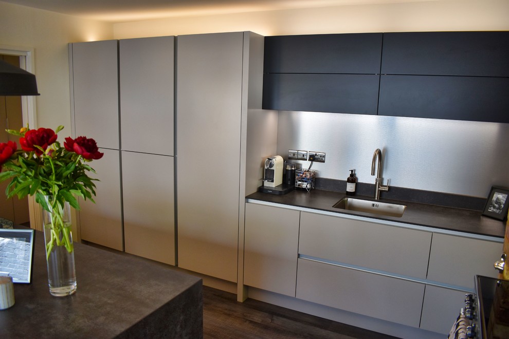 This is an example of a large contemporary galley eat-in kitchen in Cambridgeshire with a triple-bowl sink, flat-panel cabinets, black appliances, dark hardwood floors, a peninsula and black benchtop.
