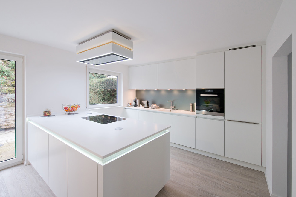 Photo of a large contemporary galley eat-in kitchen in Stuttgart with an integrated sink, flat-panel cabinets, white cabinets, solid surface benchtops, blue splashback, glass sheet splashback, light hardwood floors, with island and stainless steel appliances.