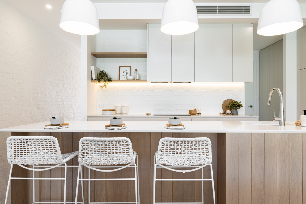 This is an example of a beach style galley kitchen in Sydney with an undermount sink, flat-panel cabinets, white cabinets, white splashback, white benchtop and with island.
