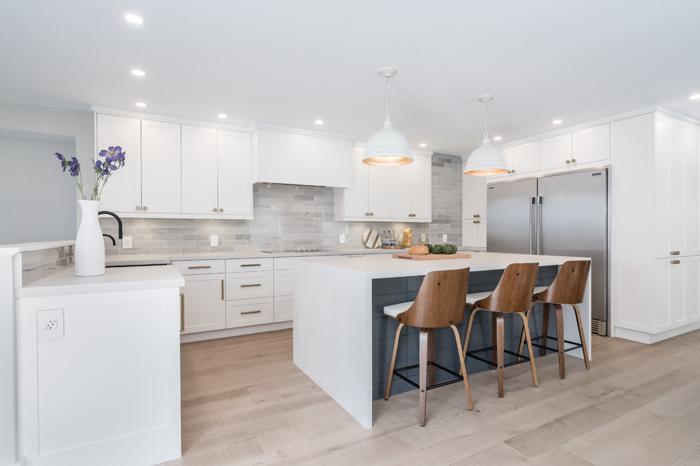 Design ideas for a transitional u-shaped kitchen in Toronto with a farmhouse sink, shaker cabinets, white cabinets, grey splashback, subway tile splashback, stainless steel appliances, light hardwood floors, with island, beige floor and white benchtop.