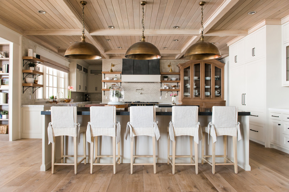 Photo of a large traditional u-shaped open plan kitchen in Salt Lake City with shaker cabinets, white cabinets, beige splashback, light hardwood floors, with island, black benchtop, a farmhouse sink, quartz benchtops, stainless steel appliances and beige floor.