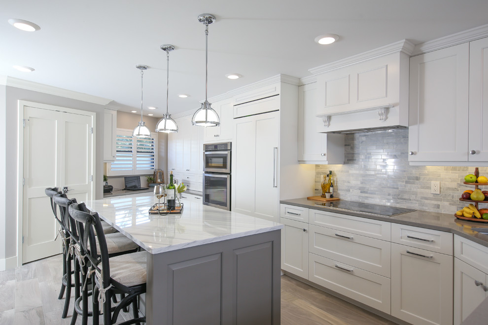 Beach style l-shaped kitchen in St Louis with recessed-panel cabinets, white cabinets, grey splashback, panelled appliances, light hardwood floors, with island, grey floor and grey benchtop.