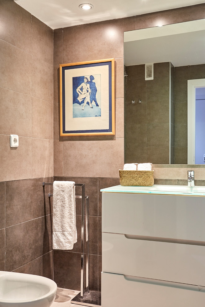 This is an example of a mid-sized modern master bathroom in Madrid with furniture-like cabinets, white cabinets, a wall-mount toilet, beige tile, ceramic tile, grey walls, ceramic floors, a trough sink, glass benchtops and beige floor.