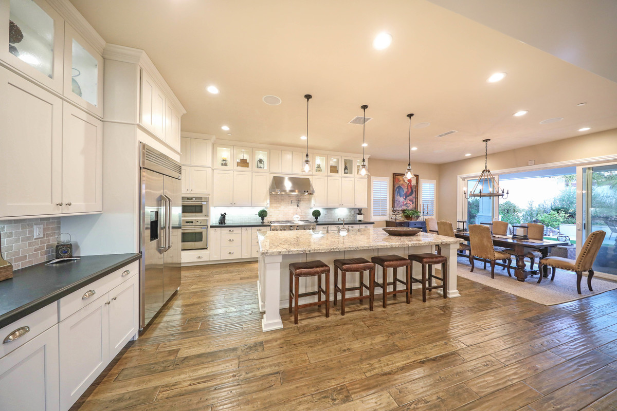 Example of a large transitional l-shaped medium tone wood floor and brown floor open concept kitchen design in Orange County with a farmhouse sink, shaker cabinets, white cabinets, quartz countertops, beige backsplash, subway tile backsplash, stainless steel appliances and an island