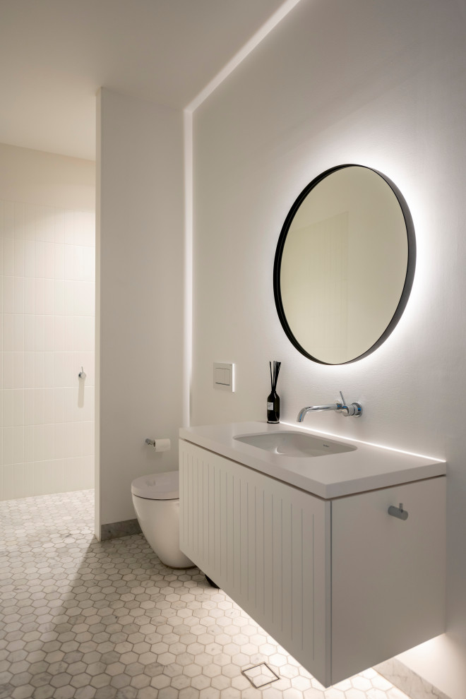 Cette photo montre un WC suspendu bord de mer de taille moyenne avec un placard à porte shaker, des portes de placard blanches, un mur blanc, un sol en marbre, un lavabo encastré, un plan de toilette en surface solide, un sol gris, un plan de toilette blanc et meuble-lavabo suspendu.