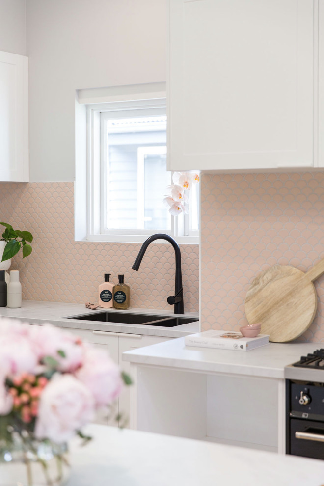 Photo of a small scandinavian l-shaped eat-in kitchen in Melbourne with an undermount sink, recessed-panel cabinets, white cabinets, quartz benchtops, pink splashback, mosaic tile splashback, black appliances, medium hardwood floors, with island and white benchtop.