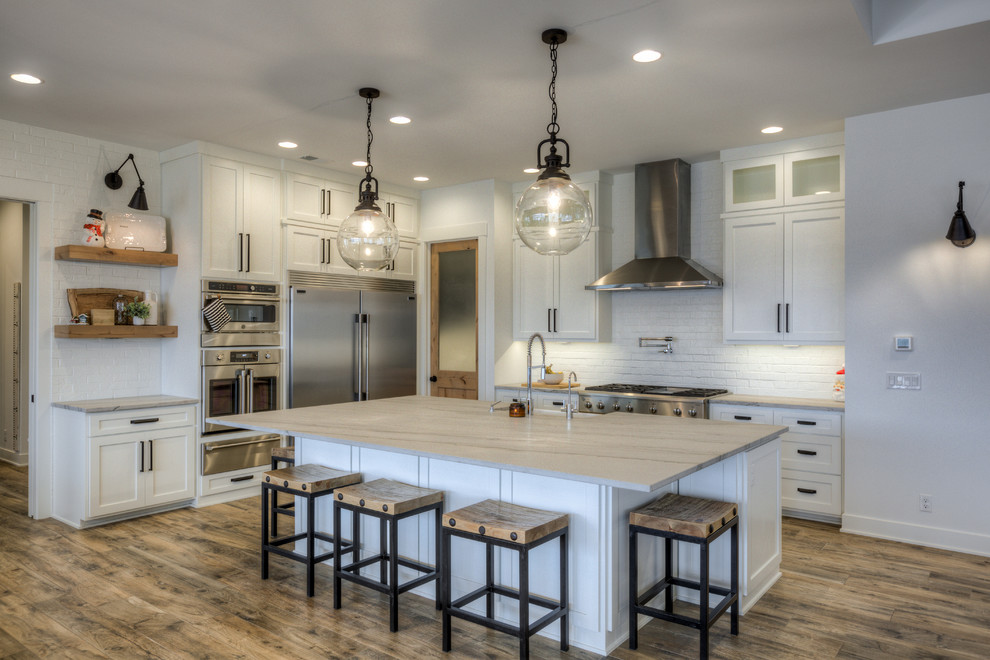Photo of a country l-shaped kitchen in Omaha with a farmhouse sink, shaker cabinets, white cabinets, white splashback, brick splashback, stainless steel appliances, dark hardwood floors, with island, brown floor and grey benchtop.