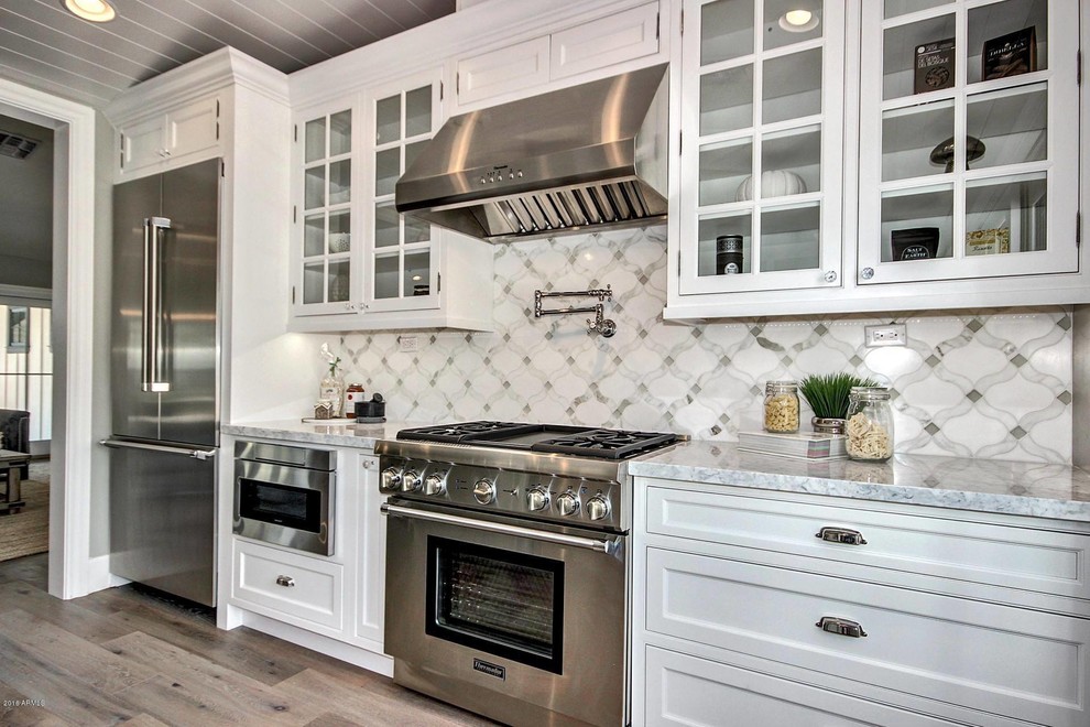 Example of a mid-sized transitional galley light wood floor eat-in kitchen design in Phoenix with a farmhouse sink, recessed-panel cabinets, white cabinets, quartz countertops, multicolored backsplash, porcelain backsplash, stainless steel appliances and an island