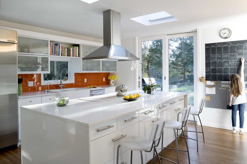 Photo of a contemporary galley kitchen in New York with a farmhouse sink, glass-front cabinets, white cabinets, quartz benchtops, orange splashback, stainless steel appliances and glass tile splashback.