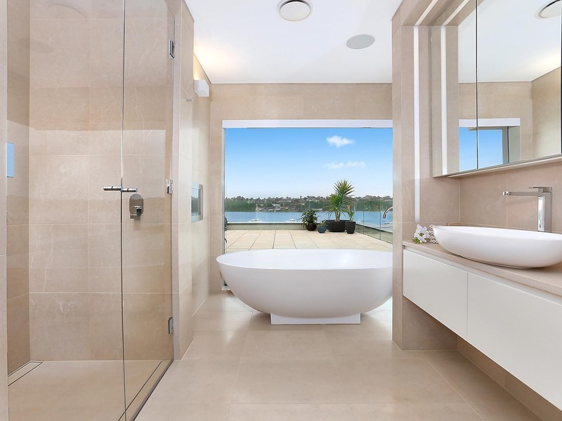 This is an example of a mid-sized contemporary bathroom in Sydney with glass-front cabinets, beige cabinets, a freestanding tub, beige tile and beige walls.