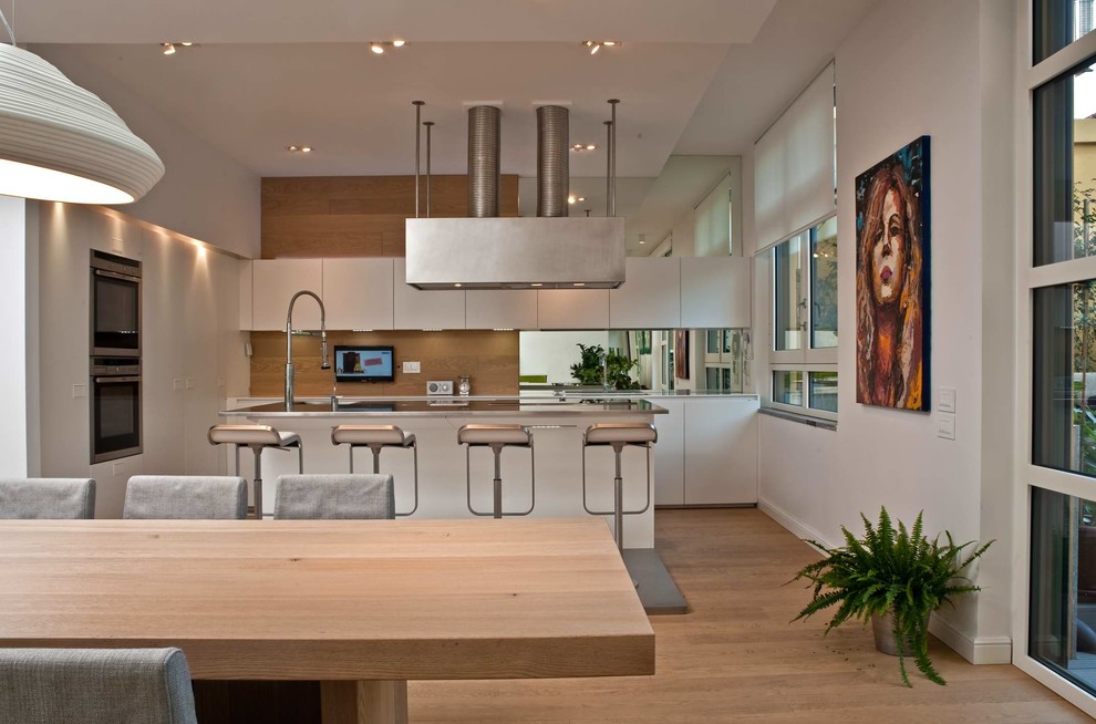 Photo of an expansive contemporary single-wall open plan kitchen in Milan with an integrated sink, flat-panel cabinets, white cabinets, stainless steel benchtops, stainless steel appliances, light hardwood floors and with island.