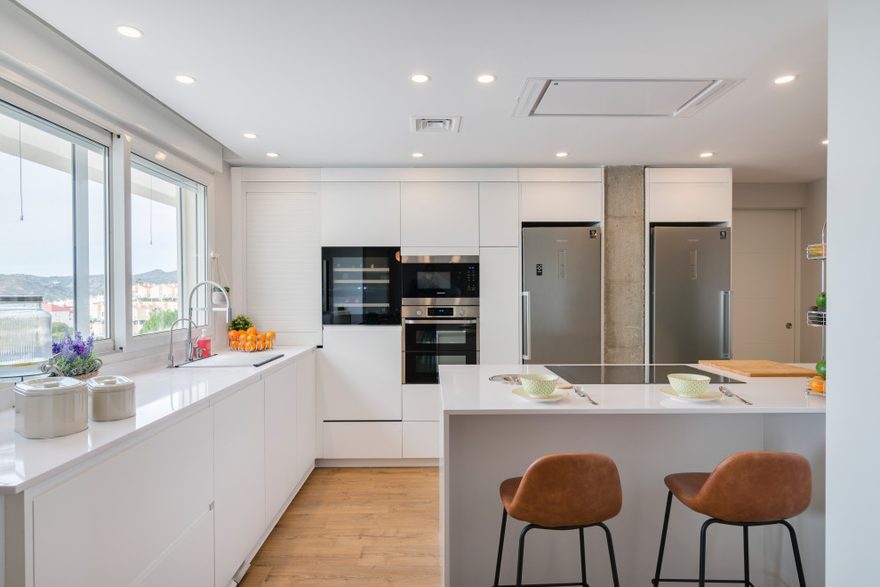Inspiration for a large contemporary l-shaped open plan kitchen in Malaga with an undermount sink, flat-panel cabinets, white cabinets, quartz benchtops, white splashback, engineered quartz splashback, stainless steel appliances, medium hardwood floors, with island, brown floor and white benchtop.