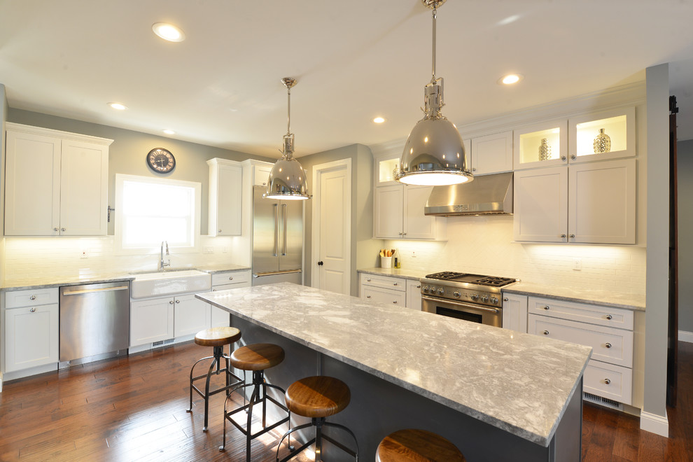 This is an example of a large transitional l-shaped eat-in kitchen in St Louis with a farmhouse sink, shaker cabinets, white cabinets, granite benchtops, white splashback, subway tile splashback, stainless steel appliances, dark hardwood floors, with island, brown floor and grey benchtop.
