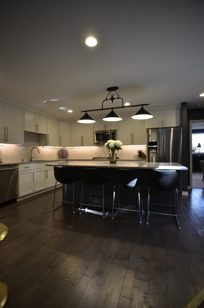 Example of a mid-sized transitional l-shaped dark wood floor and brown floor open concept kitchen design in Minneapolis with an undermount sink, flat-panel cabinets, white cabinets, granite countertops, white backsplash, ceramic backsplash, stainless steel appliances and an island