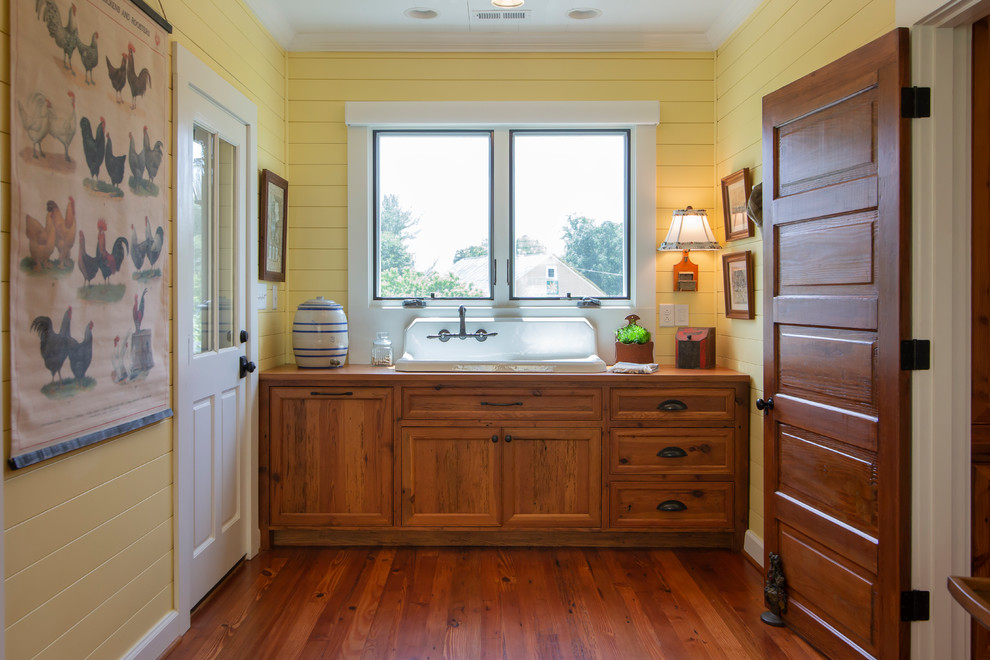 Photo of a mid-sized country single-wall utility room in Charlotte with a farmhouse sink, medium wood cabinets, wood benchtops, yellow walls, medium hardwood floors, a concealed washer and dryer and recessed-panel cabinets.