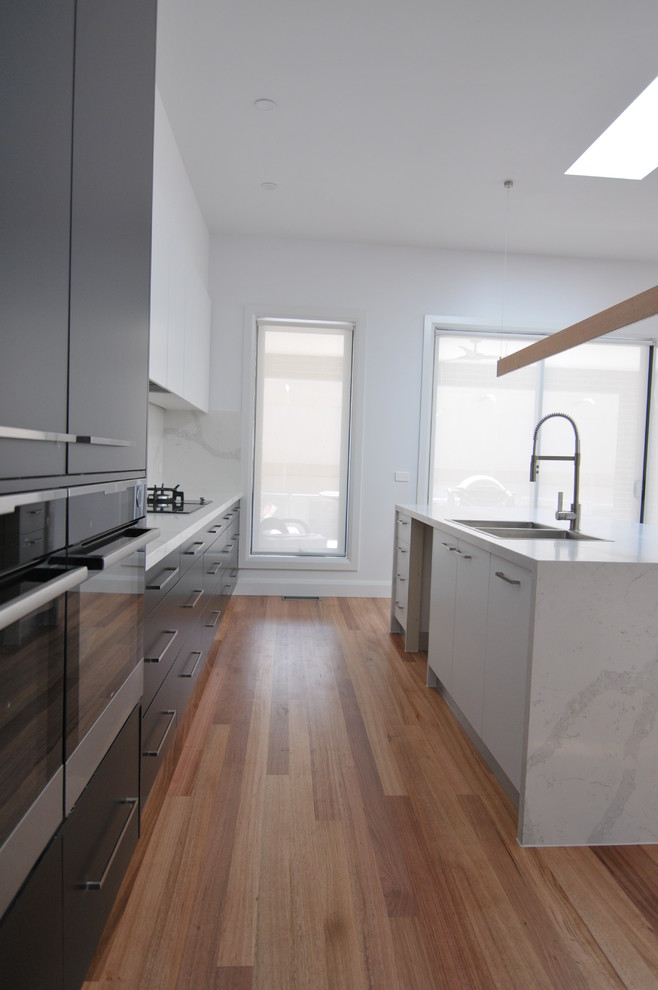 Large contemporary galley kitchen pantry in Melbourne with a double-bowl sink, flat-panel cabinets, black cabinets, solid surface benchtops, white splashback, stone slab splashback, black appliances, medium hardwood floors, with island and brown floor.