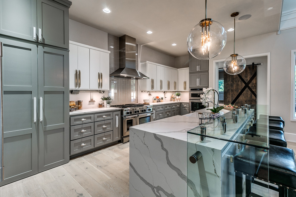 This is an example of a mid-sized transitional galley open plan kitchen in Oklahoma City with a farmhouse sink, shaker cabinets, grey cabinets, grey splashback, stainless steel appliances, light hardwood floors, with island, marble benchtops, stone tile splashback and beige floor.